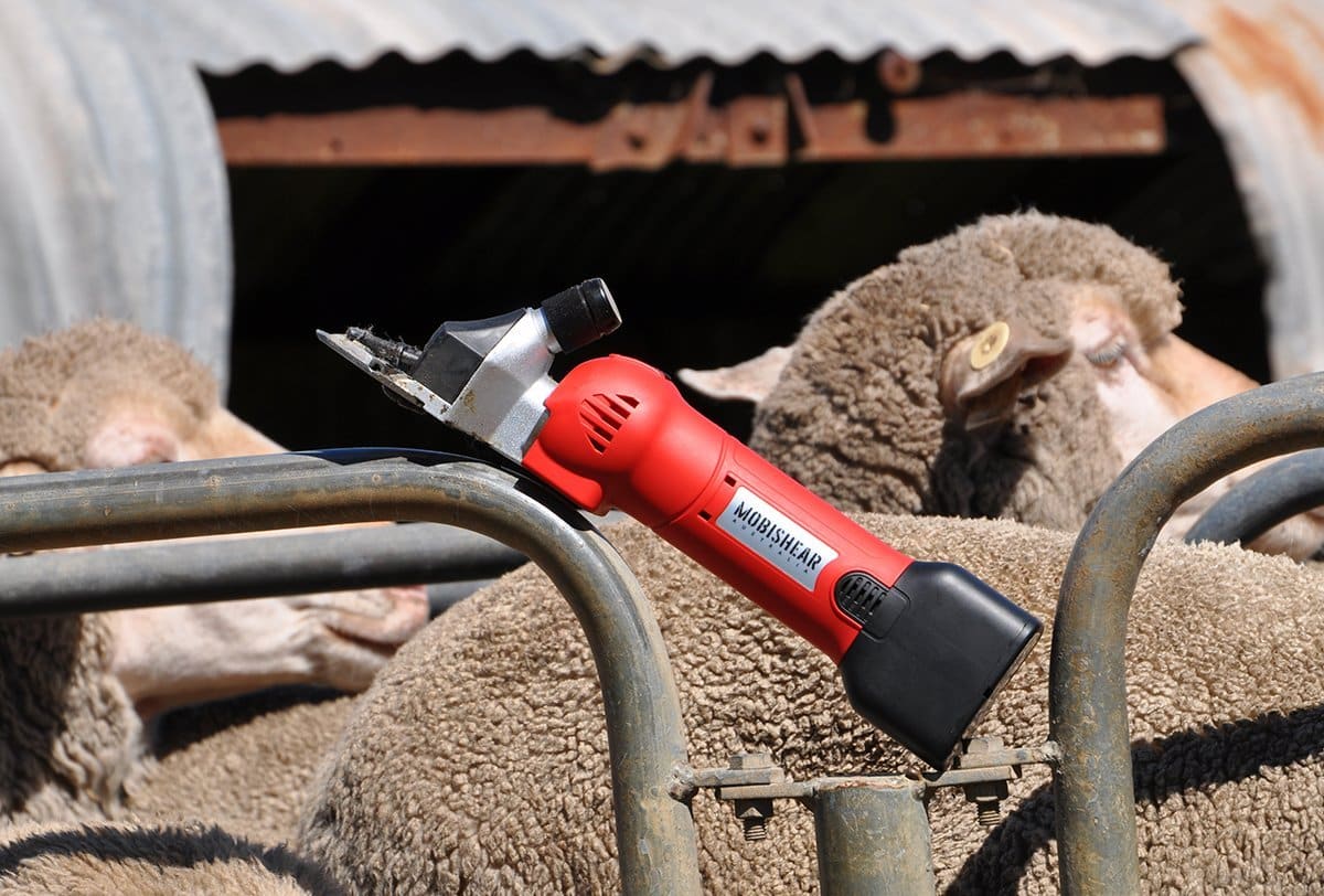 Red Mobishear Cordless Sheep Shearing Handpiece leaning on rail in sheep yards with sheep in background